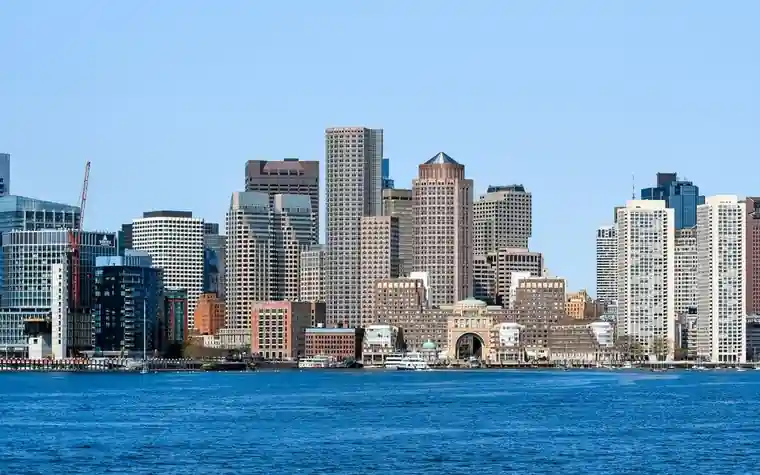 Boston skyline of glass and stone office towers across a bright blue stretch of harbor water under a clear sky