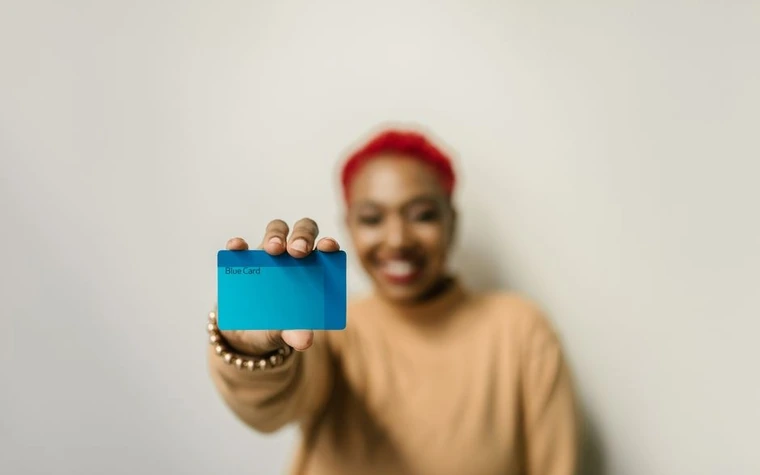 smiling woman with short red hair holding up a blue plastic card toward the camera against a plain wall