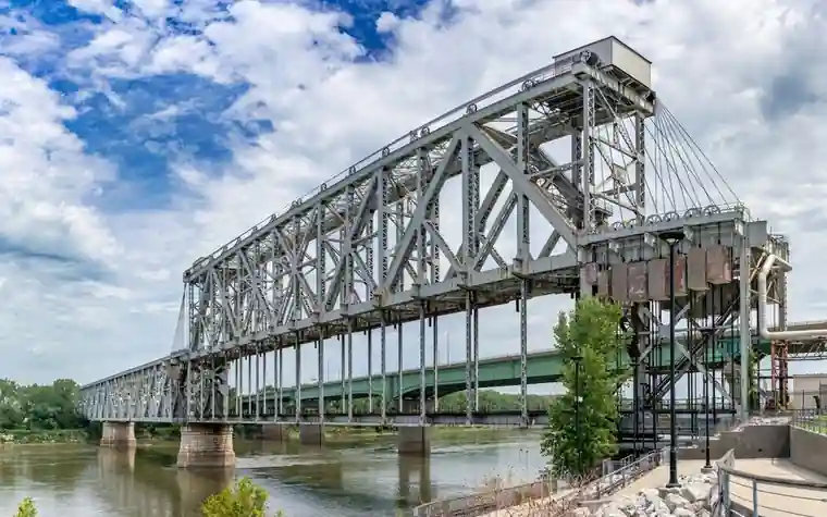 large steel truss railroad bridge spanning a wide brown river with green trees on the bank under a cloudy blue sky