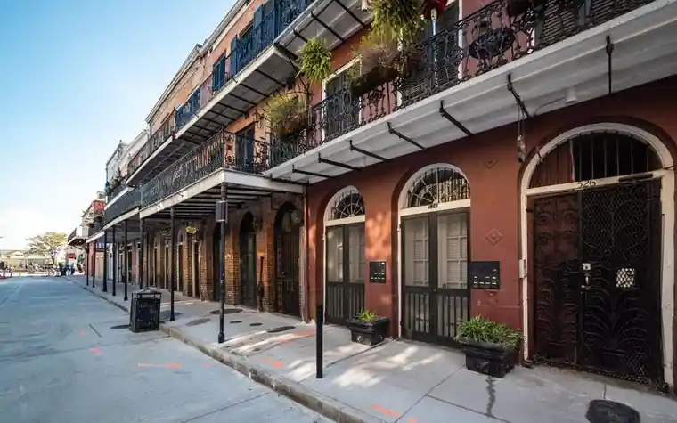 narrow New Orleans street lined with brick buildings and wrought-iron balconies with plants, shadows across the pavement on a clear day