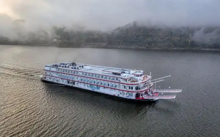 white multi-deck riverboat cruising on a wide gray river under low cloud, wooded hills fading into mist behind it