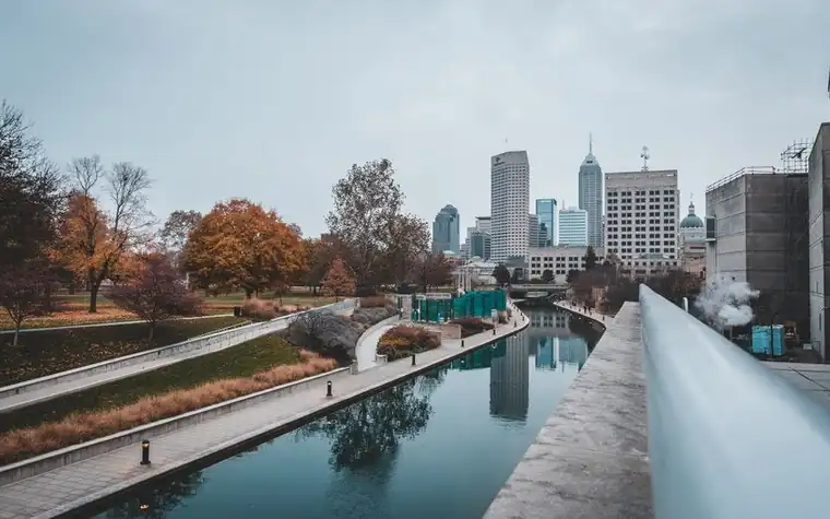 canal walkway with autumn trees beside still water and downtown Indianapolis office towers in the background
