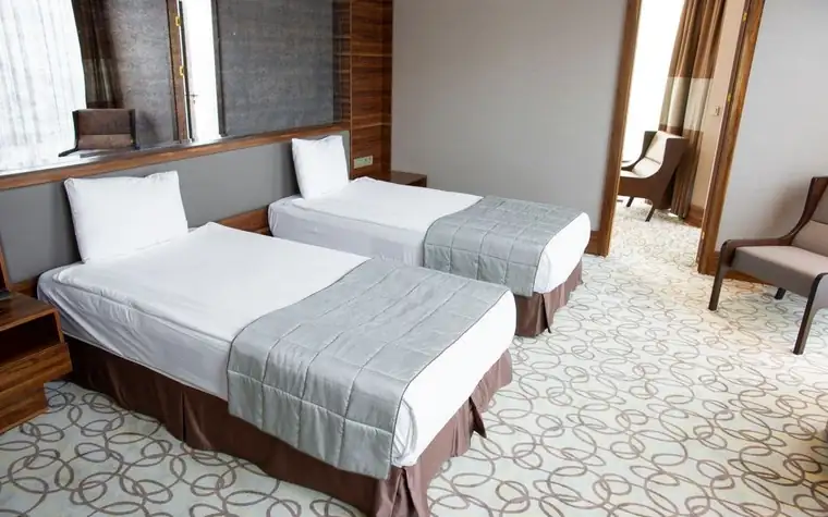 hotel room with two made twin beds, grey bed runners, a patterned carpet and an armchair by the window