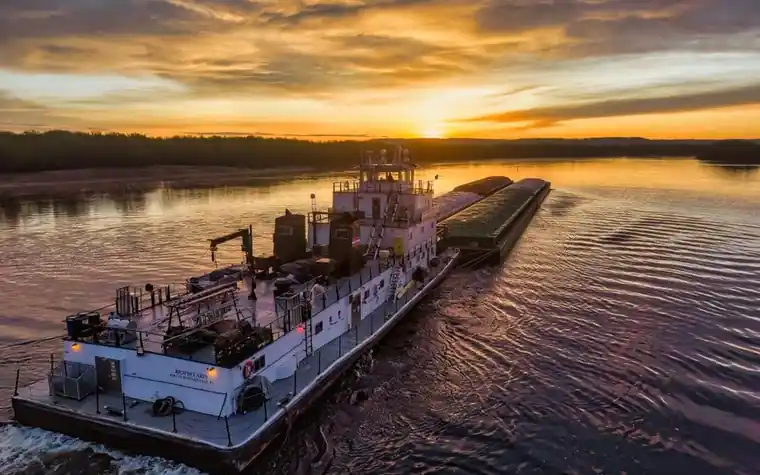towboat pushing a long barge down a wide river at sunset under a cloudy orange sky