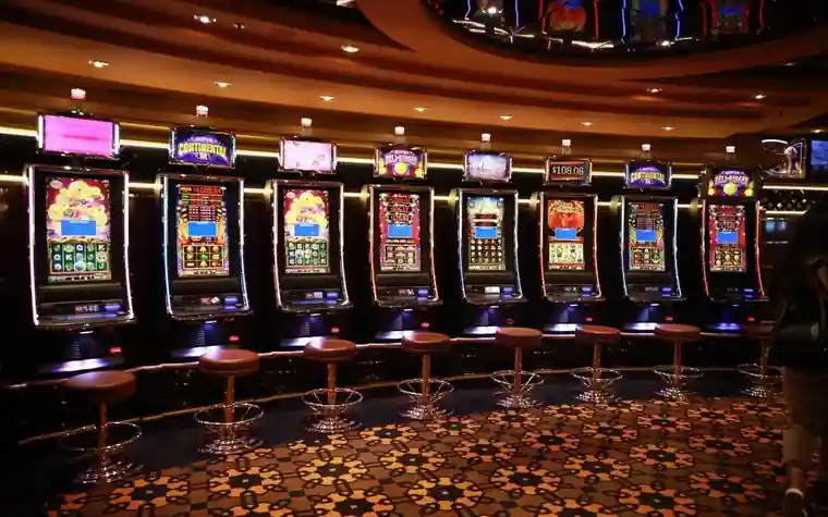 Row of illuminated slot machines with empty leather stools along a curved casino bar